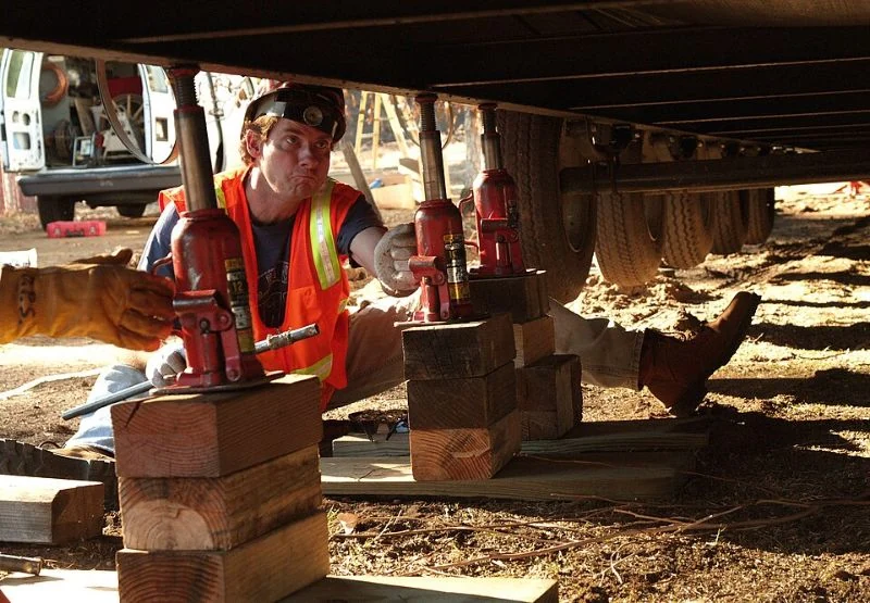 A worker installing supports beneath a mobile home, with chassis members and wheels visible.