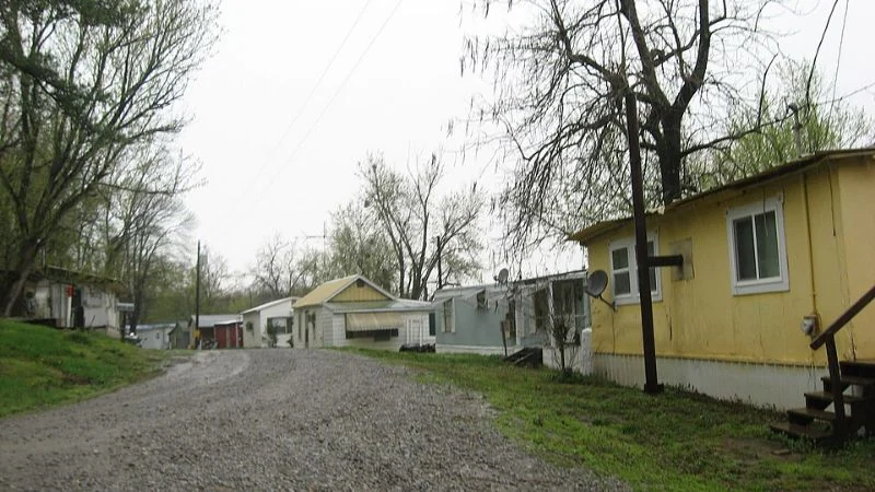 An unpaved residential lane beside mobile homes and trees under an overcast sky.